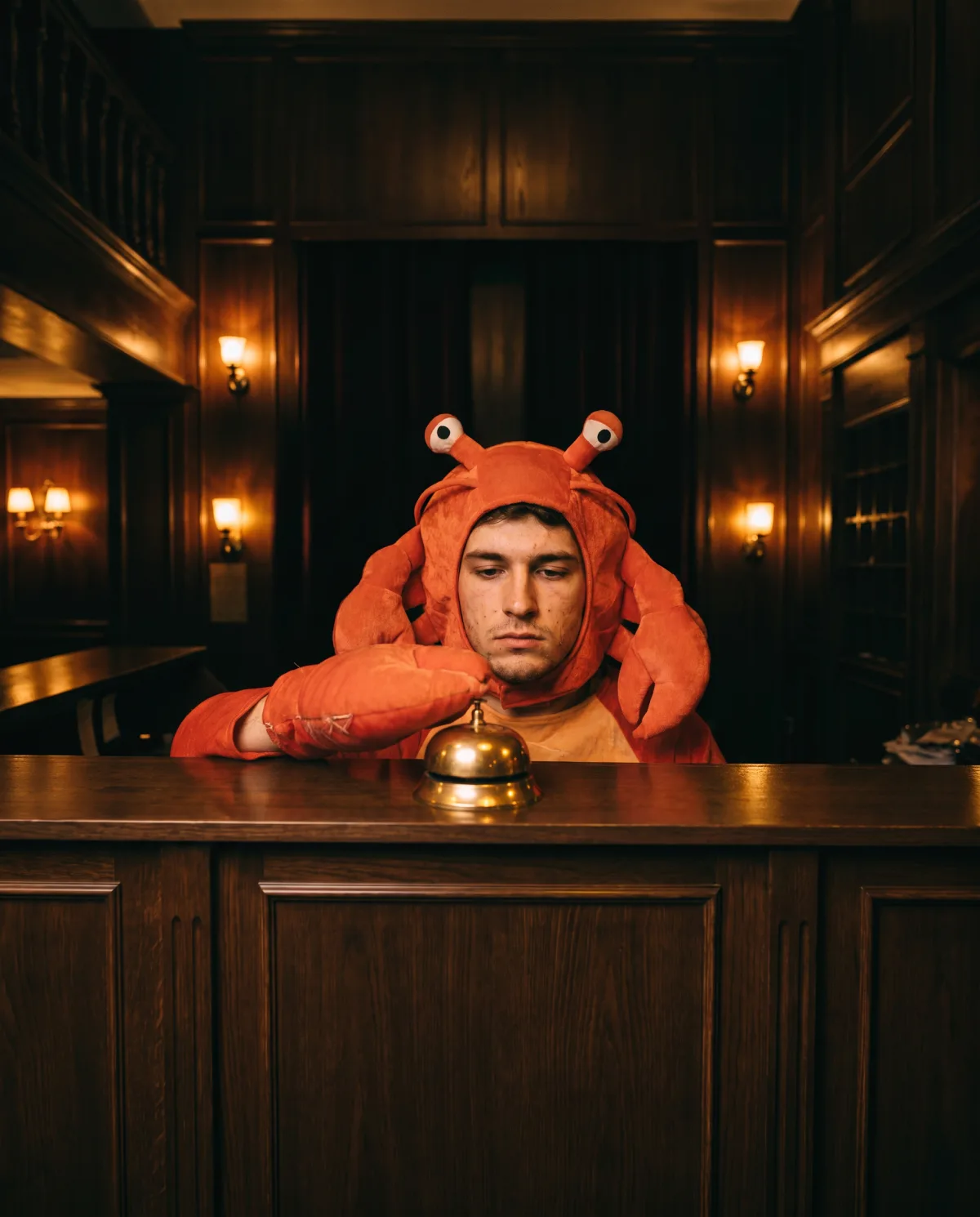 A man in a crab costume at a hotel desk, pressing its brass bell with a padded claw